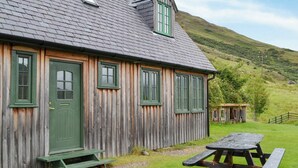 En træhytte med grøn dør og vinduer, et picnicbord og et lille skur i en græsmark med en bakke i baggrunden.