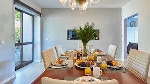A dining area with a wooden table set for breakfast, a chandelier, and a large window. - Central Holiday Home Pool by Portugal Portfolio (Cascais)