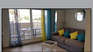 Un salón con un sofá marrón, una mesita blanca pequeña y vistas a un espacio exterior con una mesa y sillas.