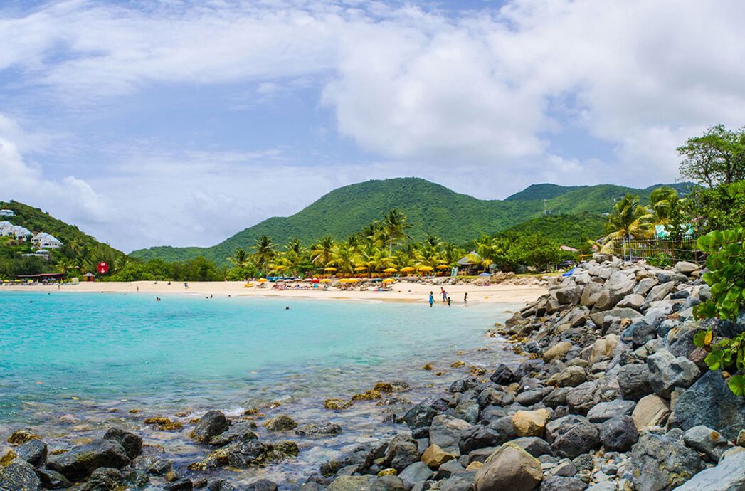A beach with clear turquoise water, rocky shoreline, and a backdrop of lush green hills.