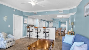 Amplia sala de estar con un sofá azul, una mesa de centro de madera y un sillón estampado. Se observa una cocina con armarios blancos y un taburete de bar al fondo.