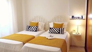 Una habitació doble amb dos llits, cadascun amb capçalera blanca, una manta groga i coixins ratllats. Hi ha una taula de nit amb una làmpada i una petita prestatgeria a sobre.