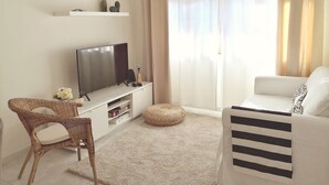 Un saló amb televisió plana, un sofà blanc, una cadira de vimet i una petita estanteria amb objectes decoratius.