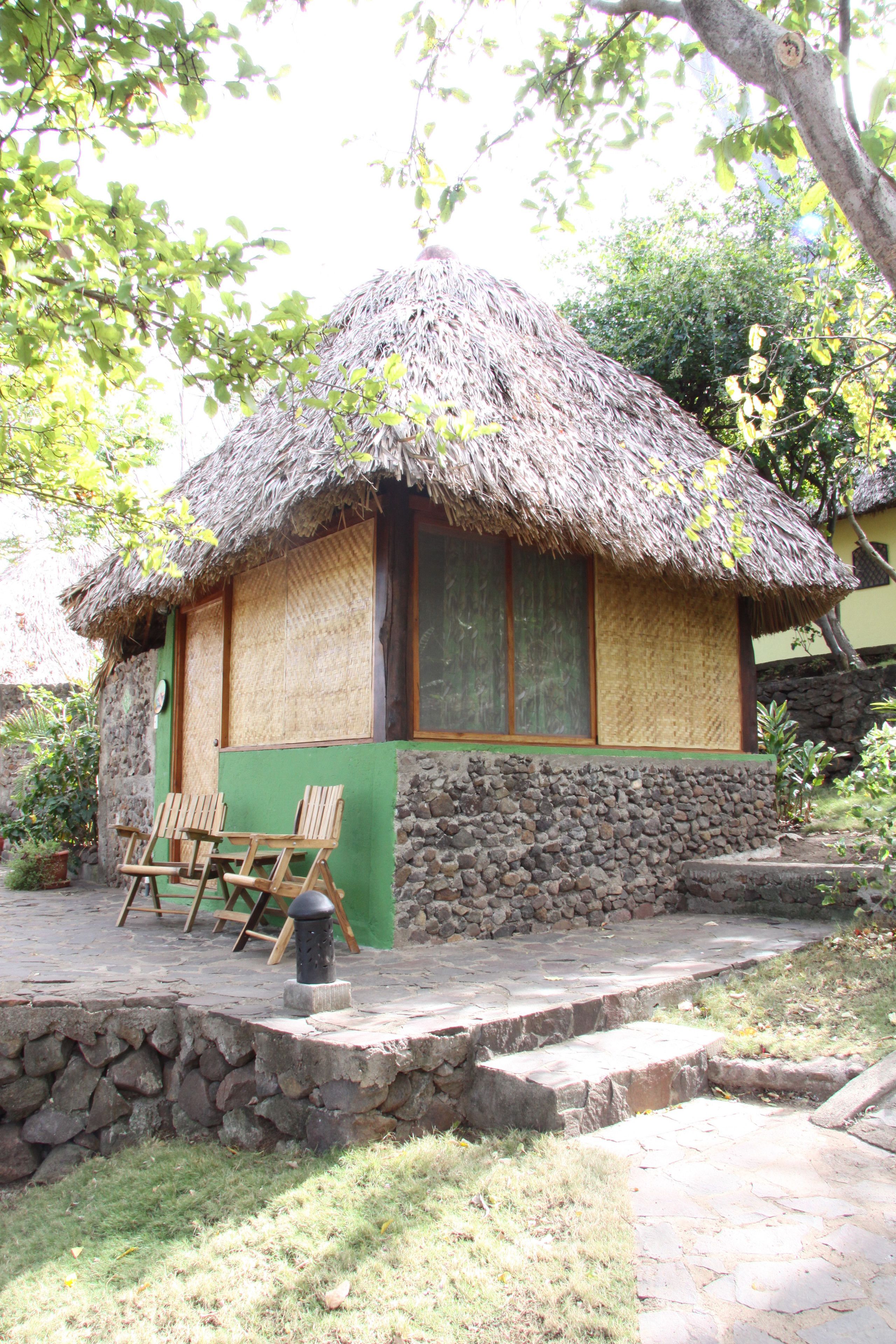 A thatched-roof hut with a stone foundation, wooden chairs outside, and a stone wall.