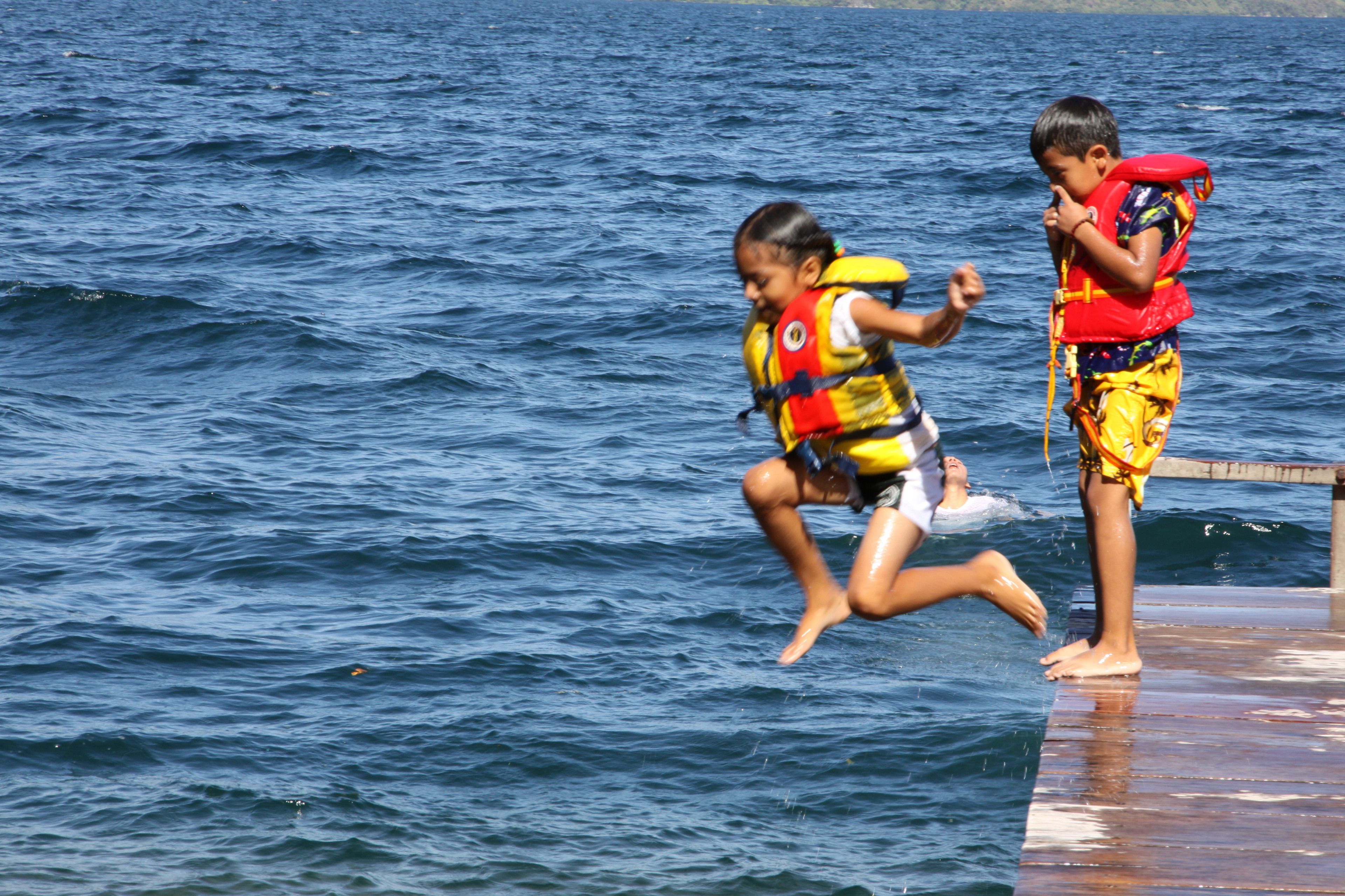 Dos niños con chalecos salvavidas saltan al agua desde un muelle.