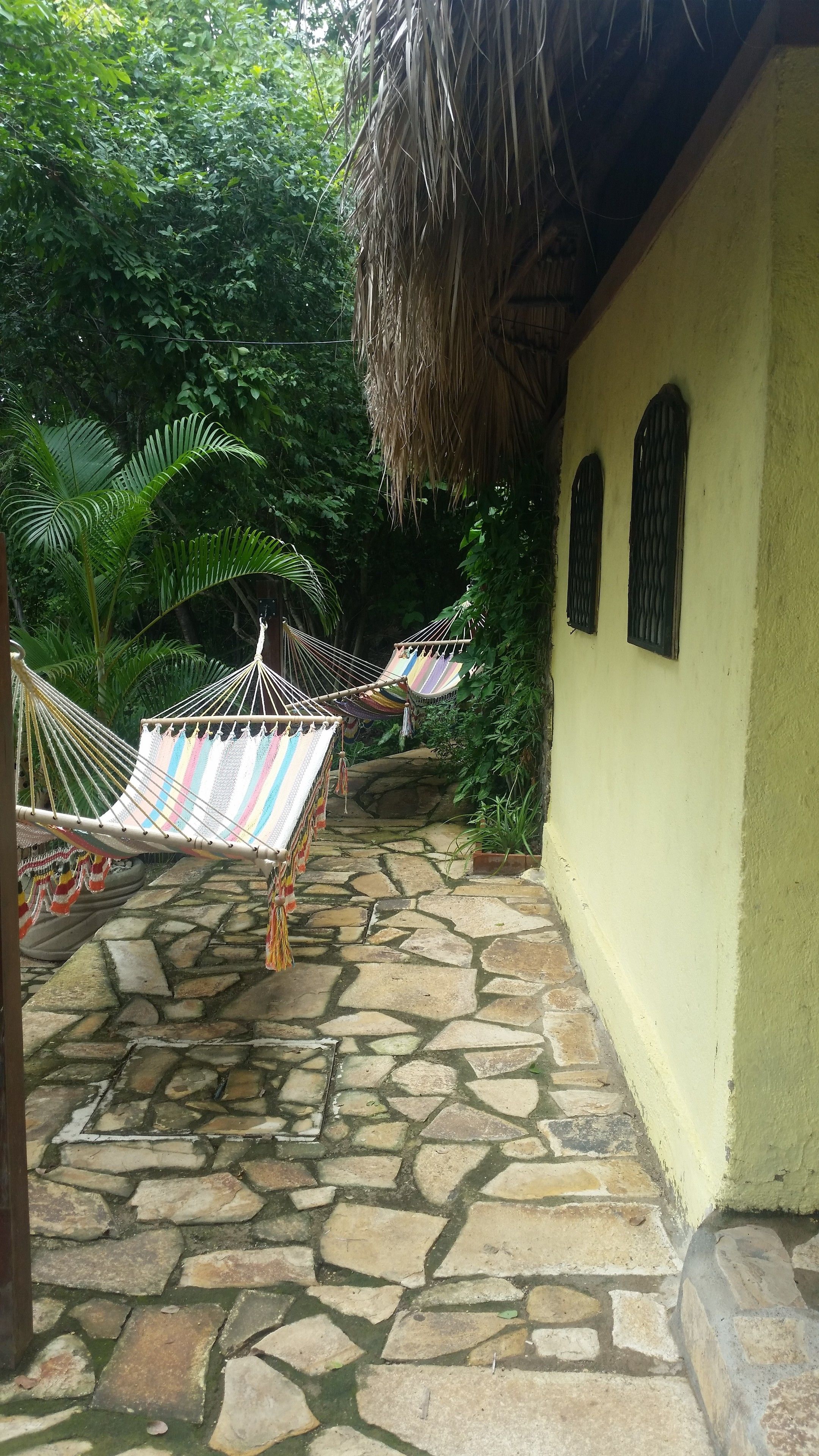 A stone-paved pathway leading to a yellow building with a thatched roof. Two hammocks are tied to the building's wall.