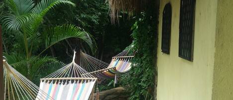A stone-paved pathway leading to a yellow building with a thatched roof. Two hammocks are tied to the building's wall.