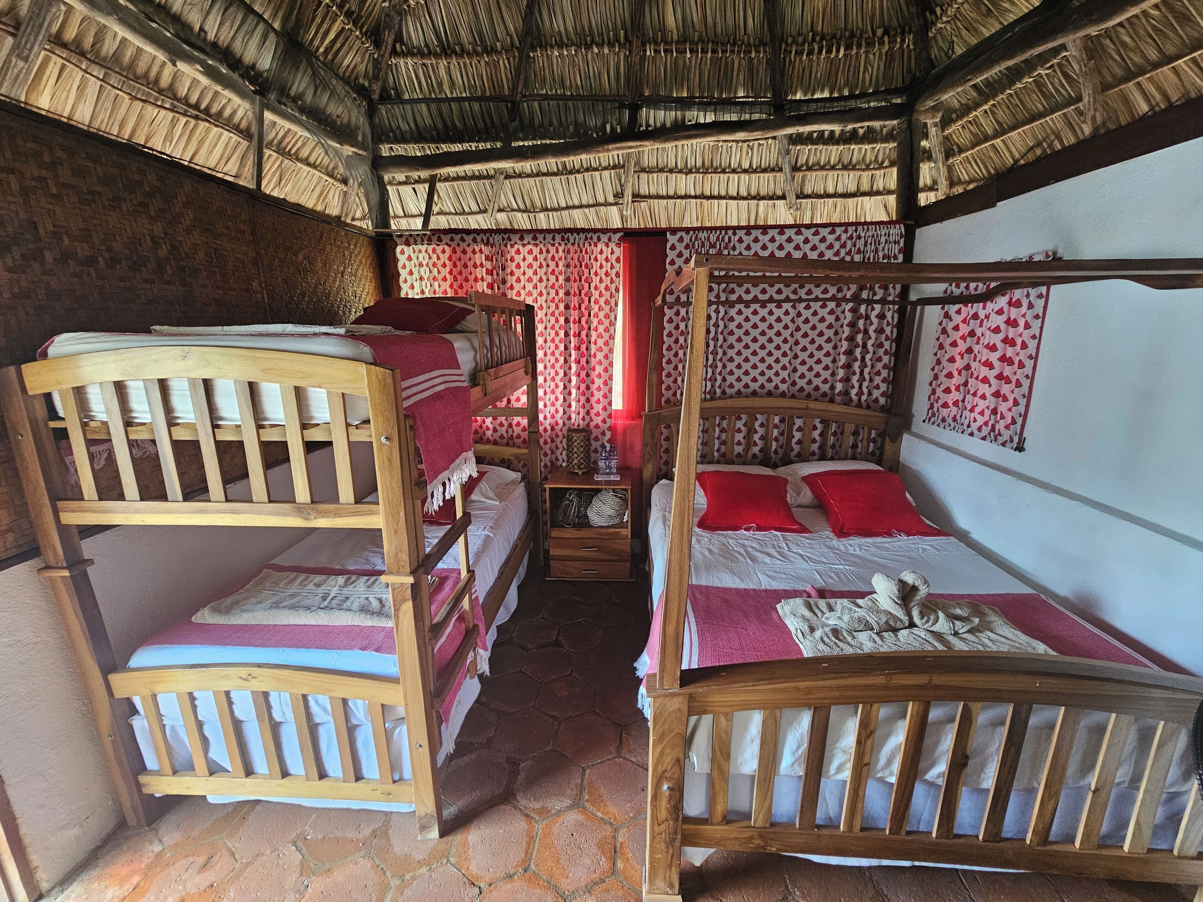 A room with bunk beds, a single bed, a patterned curtain, and a thatched roof.