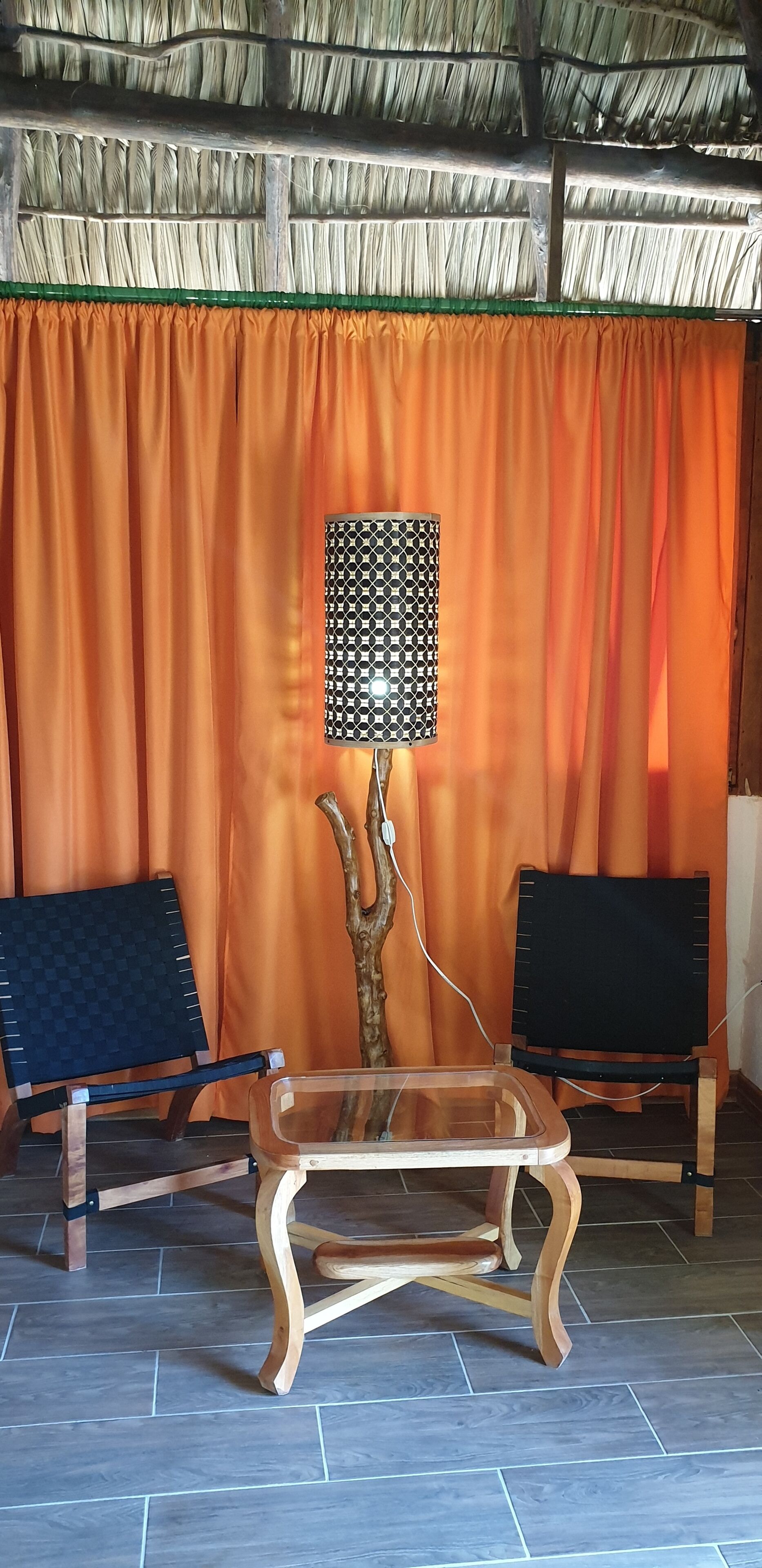 A room with a thatched roof, orange curtains, two wooden chairs, a table with a transparent top, and a decorative lamp.