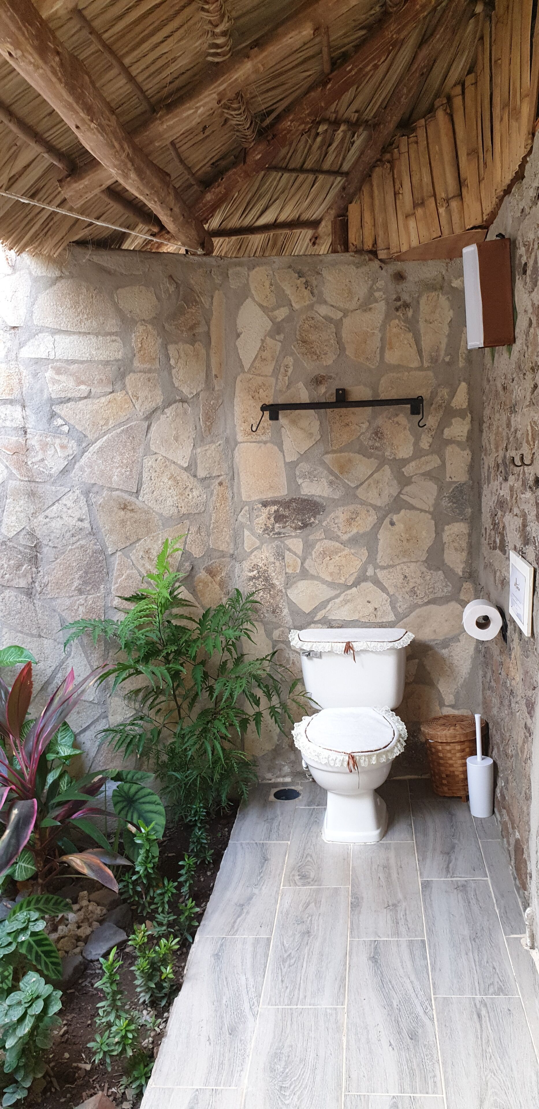 A rustic bathroom with a stone wall, a wooden ceiling, and a toilet with a decorative cover.