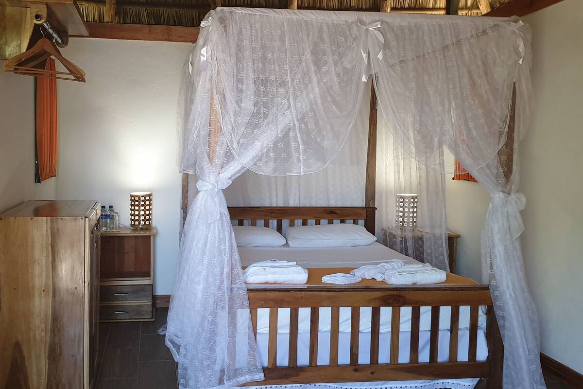 A wooden bed with a canopy, a wooden wardrobe, a nightstand with bottles, and a wall-mounted lamp.