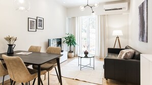 A modern living room with a dark sofa, a dining table with chairs, and a large window with curtains. - UPSTREET Peaceful 2BD Apartment in Kolonaki (Athens)