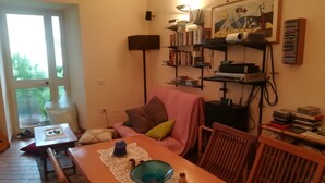 Un salon chaleureux comprenant une table à manger, des chaises, un canapé avec des coussins, une bibliothèque et une fenêtre donnant sur un paysage verdoyant.