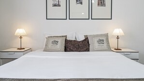 Una cama bien hecha con dos almohadas, cada una con etiqueta, flanqueada por dos mesitas de noche con lámparas. Tres cuadros enmarcados adornan la pared sobre la cama.