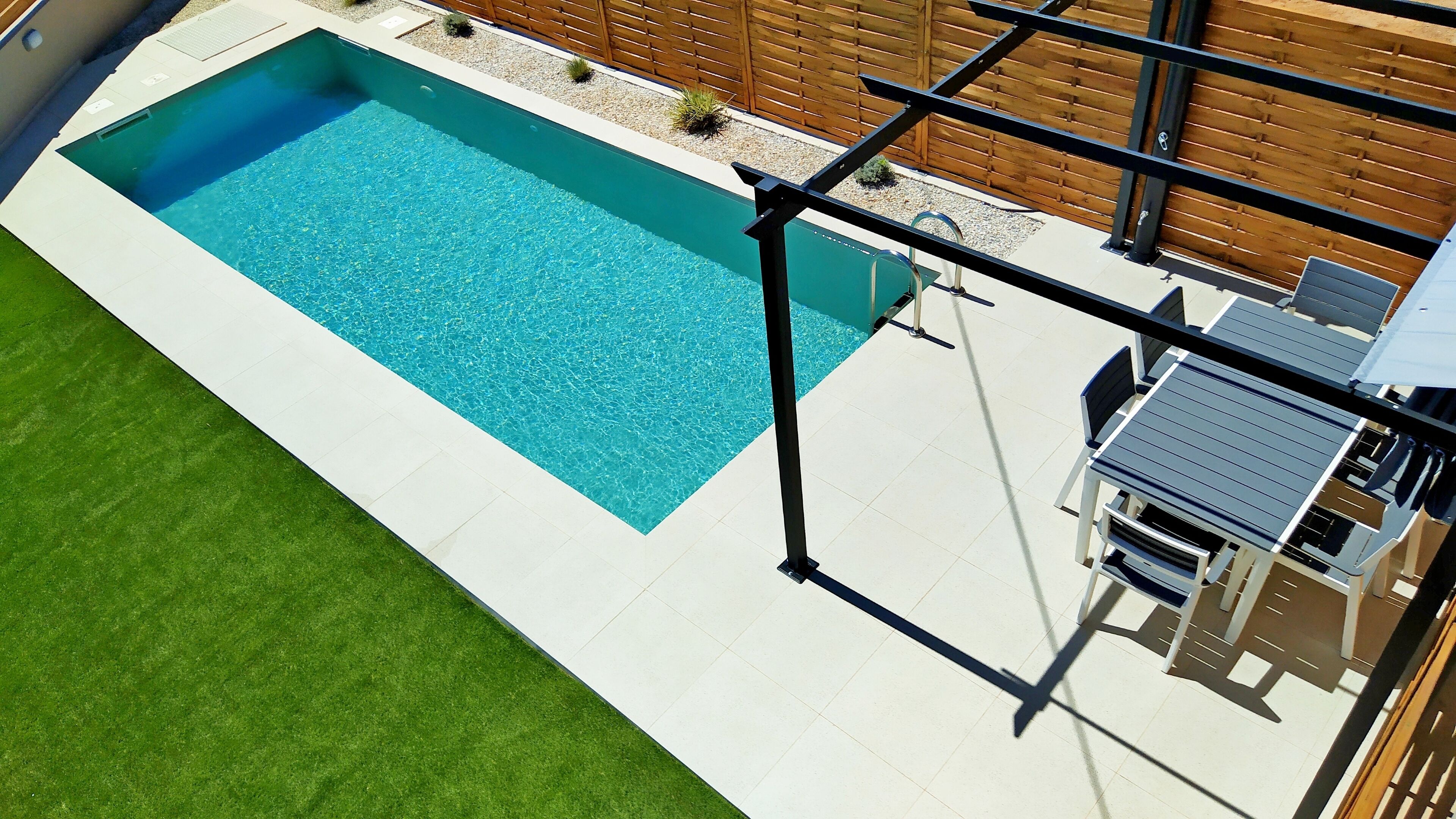 Ein rechteckiger Swimmingpool mit klarem, blauem Wasser, umgeben von einer weißen Fliesenveranda und einem grünen Rasen.