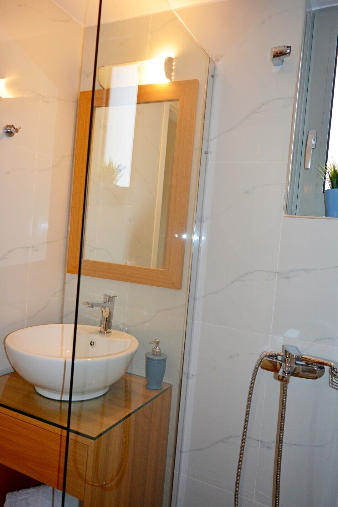 Une salle de bain dotée d’un lavabo entouré de parois en verre, d’un meuble-lavabo en bois, d’un miroir avec un cadre en bois et d’une douche avec un pommeau de douche à main.