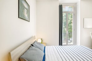 Un dormitorio con cama, mesita de noche, ventana con cortinas y un cuadro en la pared.
