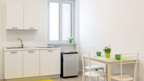 Et kompakt køkken med hvide skabe, et lille køleskab, et vindue og et spiseområde med stole og et bord.