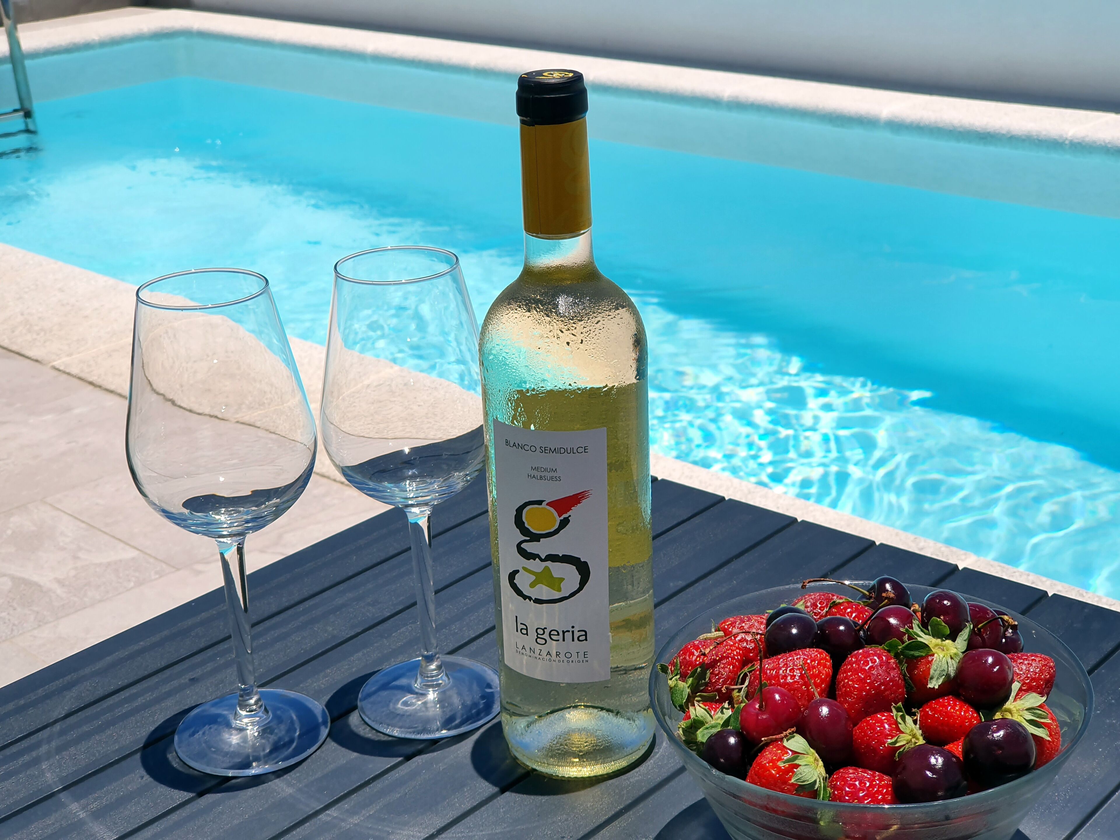 A bottle of Blanco Semidulce wine, two empty wine glasses, and a bowl of strawberries and cherries by a pool.