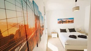Una habitación de hotel con una cama, una mesita de noche, un cuadro colgado de un puente, un cuadro de un paisaje urbano y una lámpara colgante.
