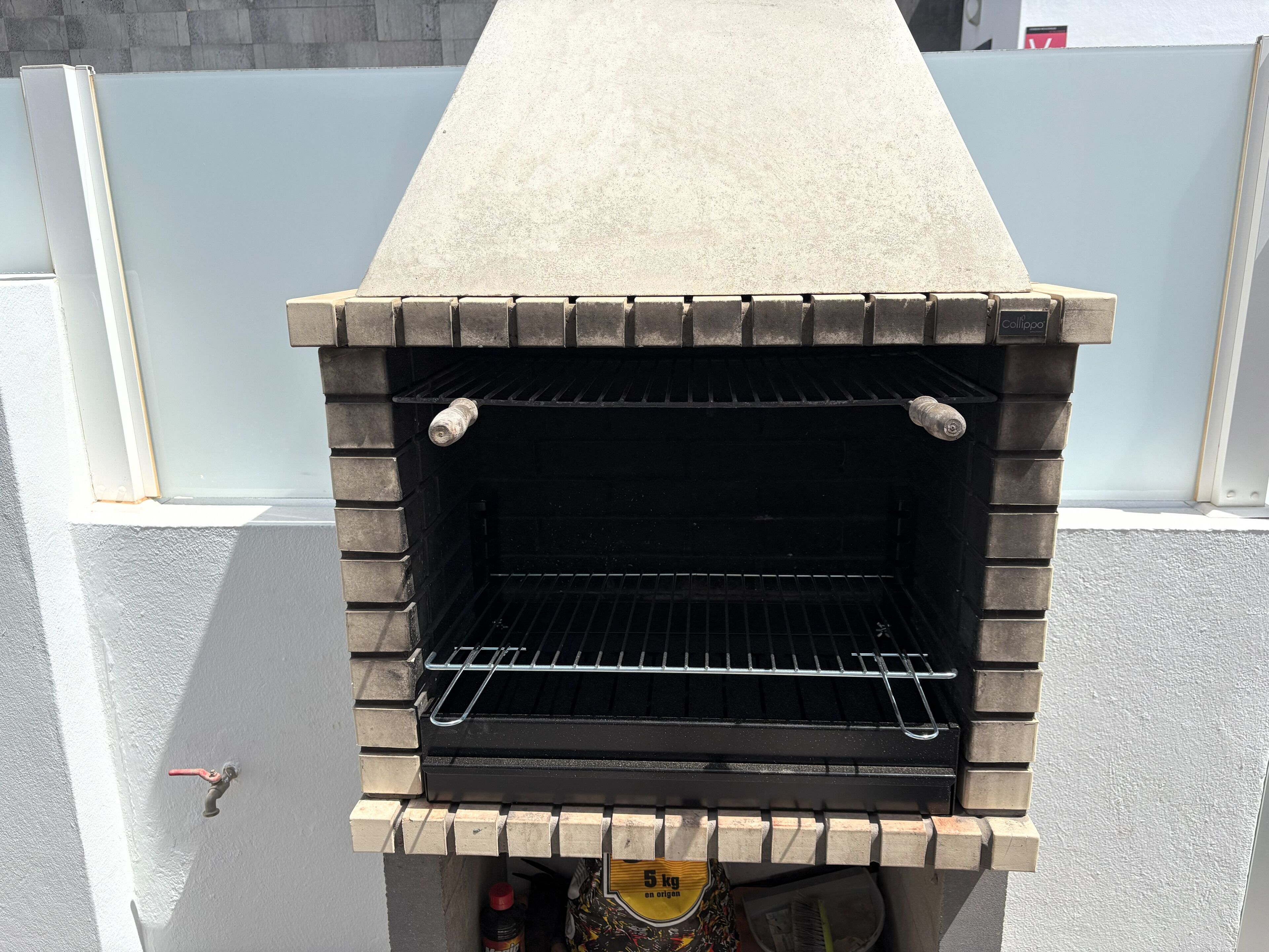 Een betonnen barbecue met een metalen rooster en een fles saus eronder.