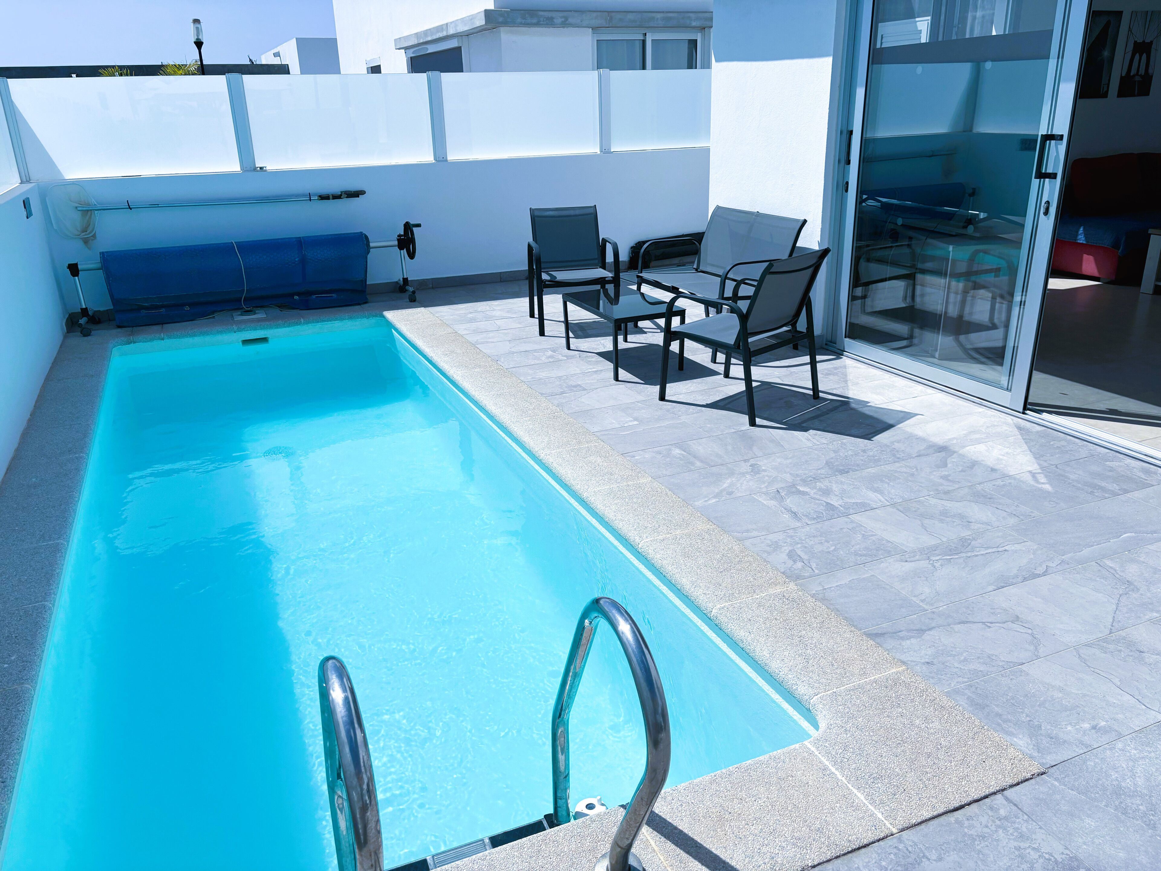 Una piscina rectangular con una barrera de vidrio y pasamanos metálicos, rodeada por un patio empedrado con mobiliario exterior.