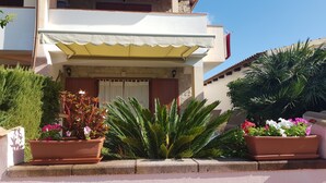 A building with a white facade, a yellow awning, and a red door. There are potted plants with pink and white flowers in front. - Home description (Viddalba)