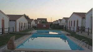 A swimming pool surrounded by white buildings with red shutters, a stone pathway, and a grassy area. - Home description (Viddalba)