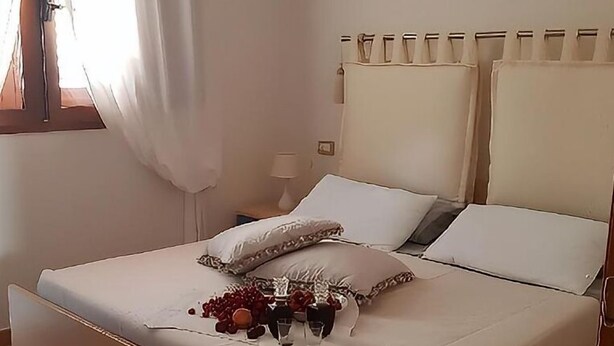 A neatly made bed with white linens and pillows, a bedside table with a lamp, and a window with curtains. - Home description (Viddalba)