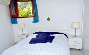 Ein Schlafzimmer mit einem Bett, zwei Nachttischen, einem Fenster mit Vorhängen und einem an der Wand befestigten Kruzifix.