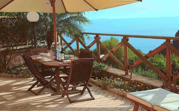 An outdoor dining area with a table and chairs, a wooden railing, and a view of the sea.
