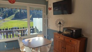 Une pièce comprenant une table et des chaises, un meuble en bois avec un four à micro-ondes, un téléviseur fixé au mur et une vue sur un paysage verdoyant par la fenêtre.