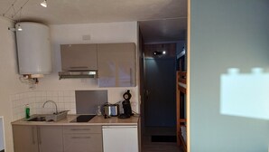 Une cuisine compacte équipée d’un évier, d’une cuisinière et d’un petit réfrigérateur. On y trouve également un petit coin repas avec une table et une chaise. Une couchette est visible dans la pièce adjacente.