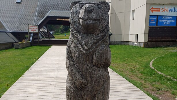 Une statue d’ours en pierre se dresse sur une promenade en bois, avec un bâtiment en arrière-plan.