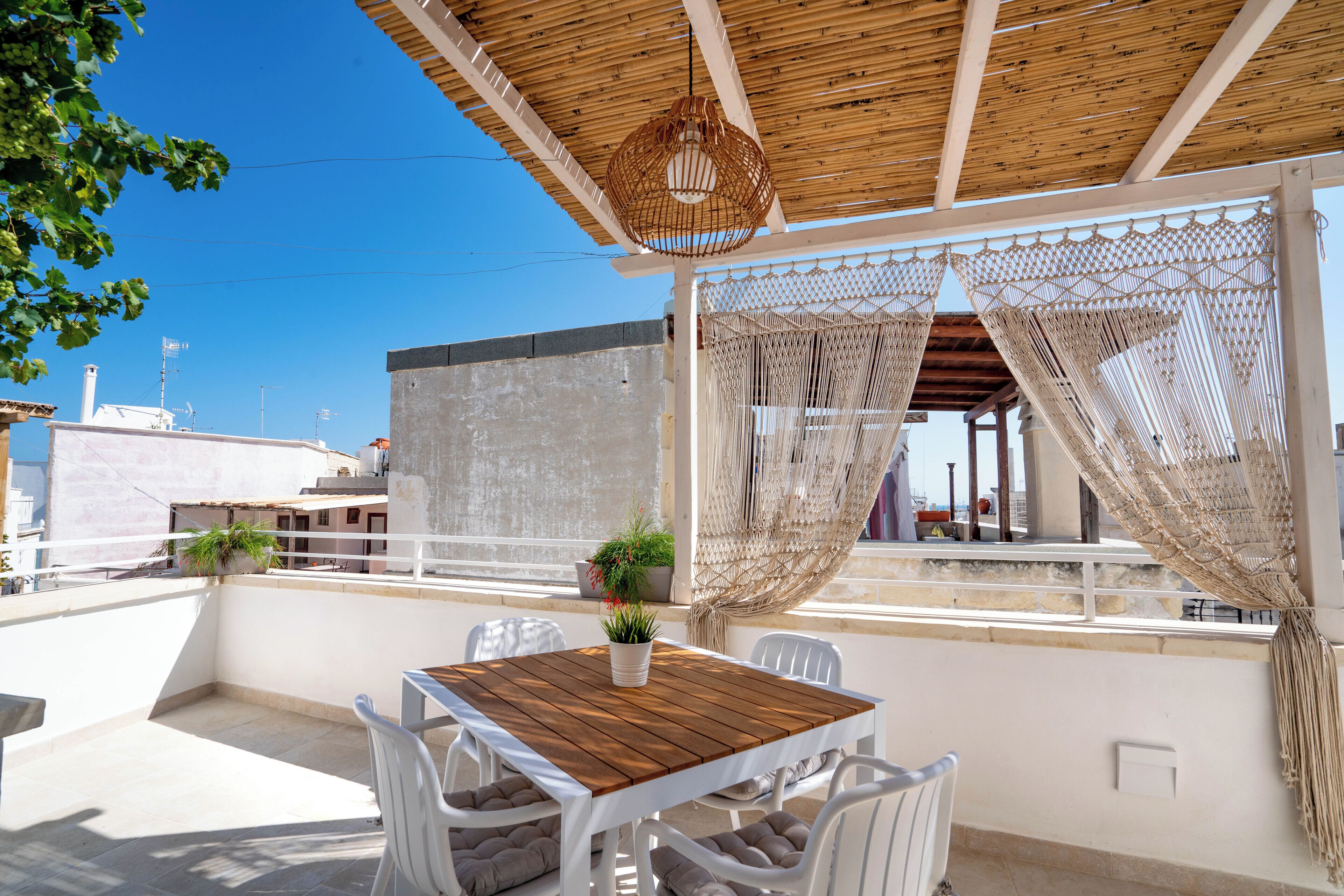 A terrace with a wooden table and white chairs, a hanging light fixture, and a view of buildings in the background.