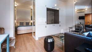 Une chambre moderne avec une cloison en verre, un lit, une petite table et un coin salon comprenant un canapé et des fauteuils.
