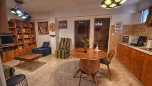 A living room with a sofa, a coffee table, and a TV. There is a dining table with chairs and a kitchen area with cabinets and appliances.