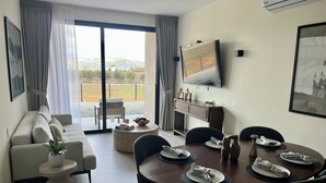 Un salón moderno con un sofá, una mesa de centro y una zona de comedor con una mesa preparada para cuatro comensales. Un televisor está montado en la pared, y se ve un jardín a través de las ventanas.