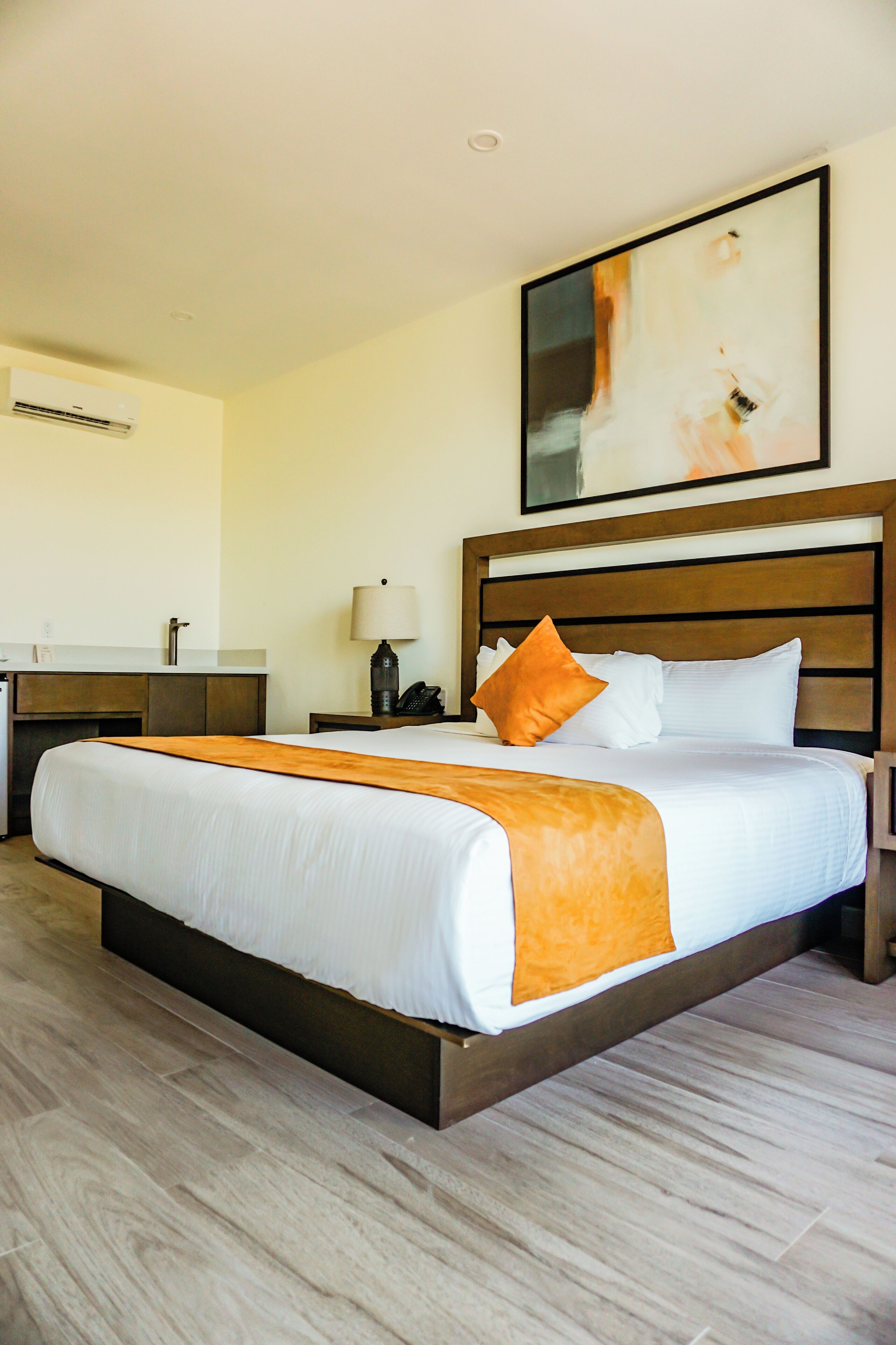 A hotel room with a large bed, a bedside table with a lamp, a nightstand, a wall-mounted air conditioner, and a painting above the bed.
