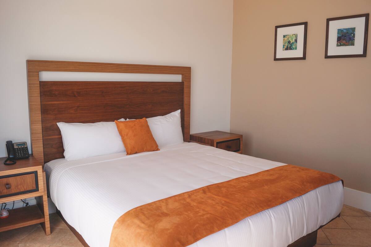 Una cama bien hecha con cabecera de madera, una mesita de noche con un teléfono y dos cuadros en la pared.