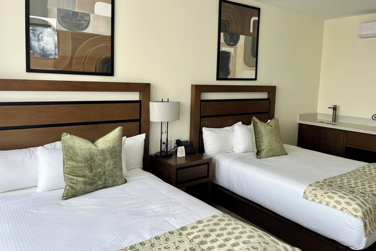 Habitación de hotel con dos camas, cada una con ropa de cama blanca y almohadas verdes, una mesita de noche con lámpara y un escritorio pequeño con teléfono.