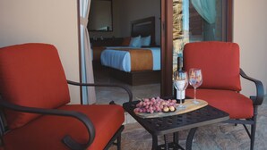 Una habitación con una cama, una mesa con una bandeja de uvas y copas de vino, y dos sillas rojas.