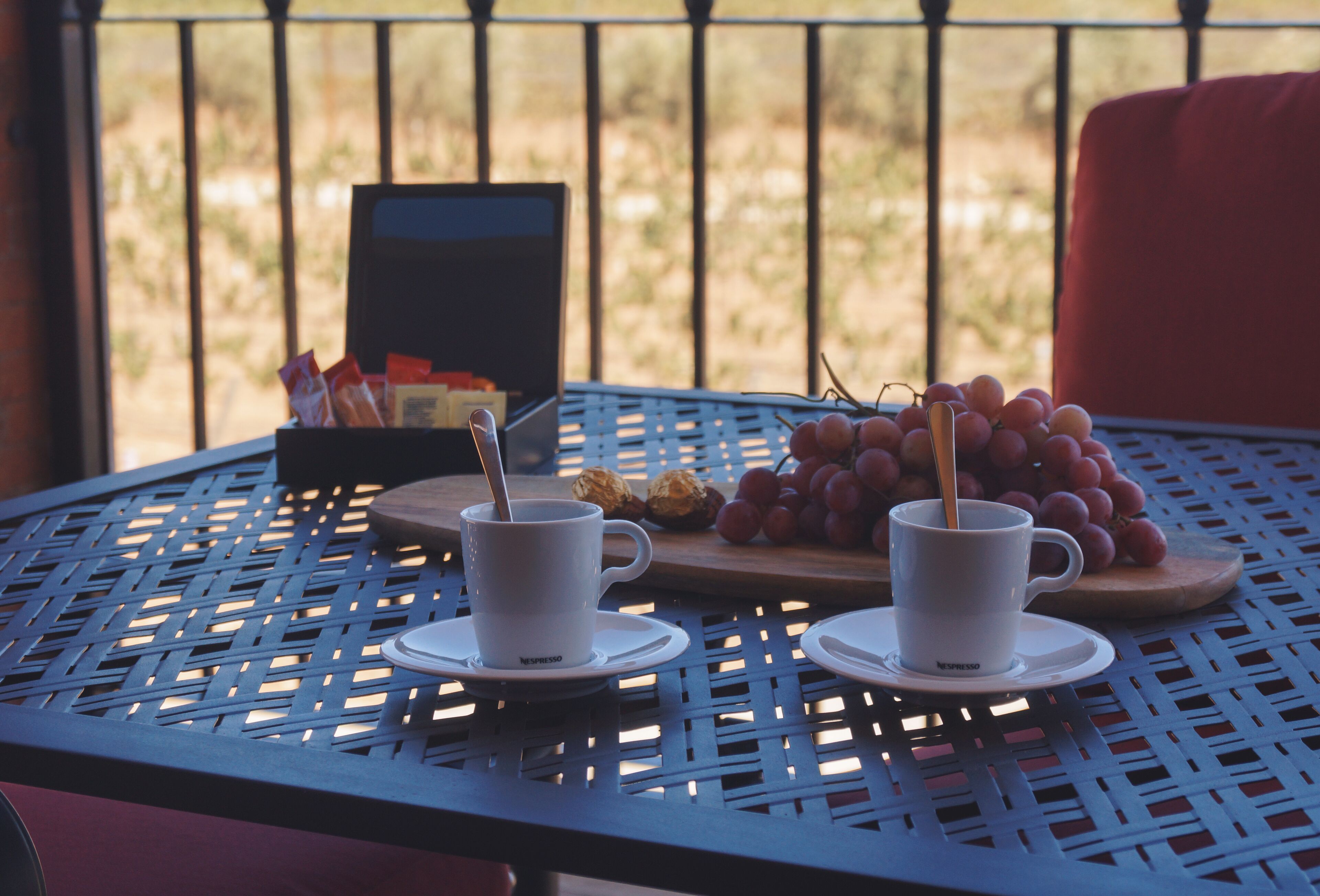 Une table avec deux tasses blanches et une assiette de raisins, placée sur un balcon doté d’une balustrade, avec une chaise rouge en arrière-plan.