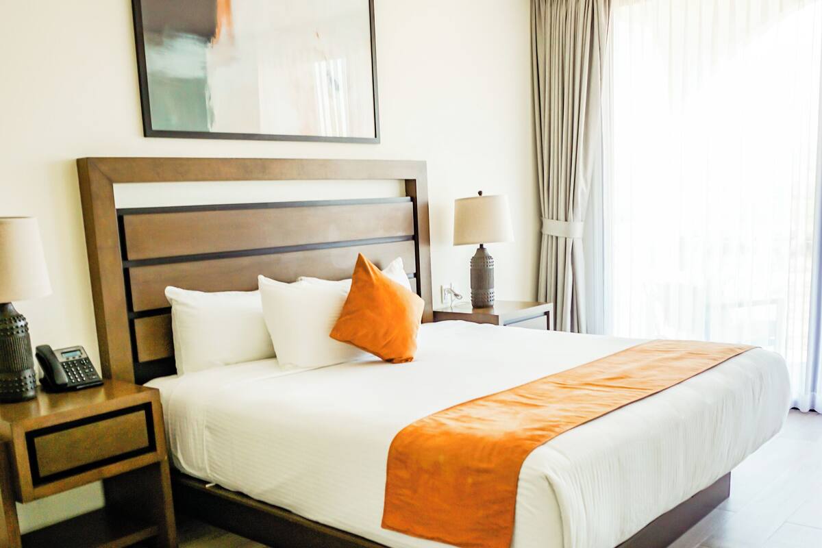 Habitación de hotel con una cama grande, cabecera de madera y vistas al exterior a través de cortinas traslúcidas.