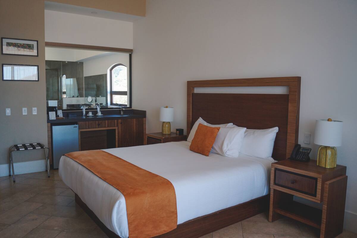 Habitación de hotel con una cama grande, una kitchenette pequeña, un espejo y una mesita de noche con lámpara.
