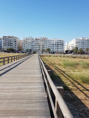 Une promenade en bois mène à une zone de plage, avec en arrière-plan des bâtiments modernes aux façades blanches.