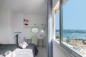 A hotel room with a bed, a desk, a chair, a round mirror, and a view of the sea.