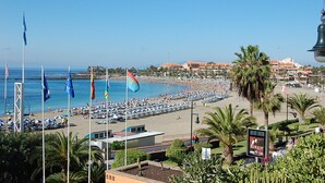 A beach with rows of sun loungers and flags, a clear blue sky, and a building in the background. - La Estrella 1A020 con vistas panorámicas al mar (Los Cristianos)