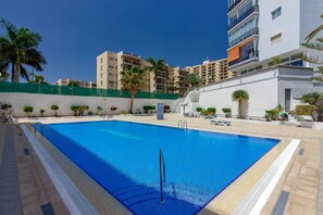 A swimming pool with a clear blue water, surrounded by a white building with balconies and palm trees.