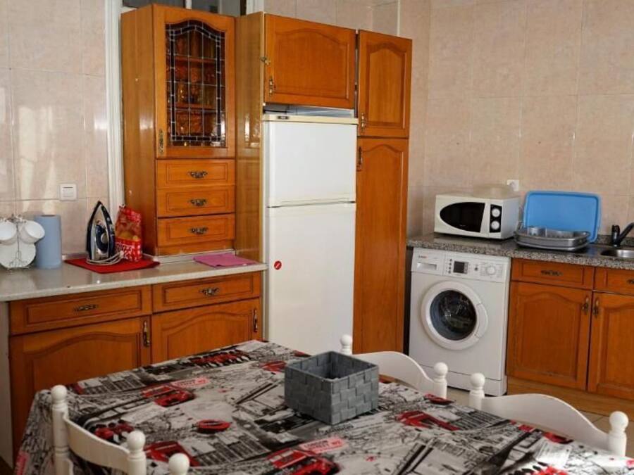 A compact kitchen with wooden cabinets, a refrigerator, a microwave, a washing machine, and a dining table with a newspaper-patterned tablecloth.